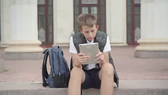 Portrait of Tired Caucasian Schoolboy Using Tablet During School Break Outdoors. Sleepy Boy Surfing alt