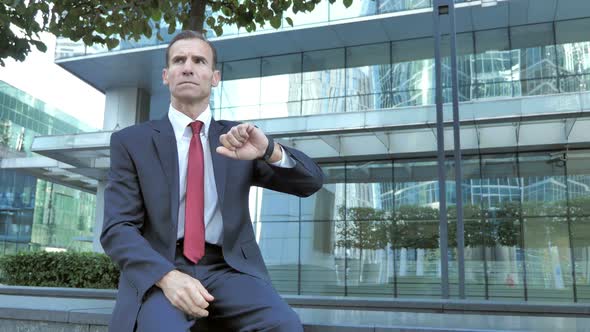Waiting Businessman Sitting Outside Office and Watching Time alt