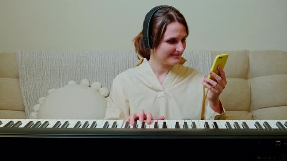 Happy woman musician with piano looking at mobile phone at home on sofa in living room