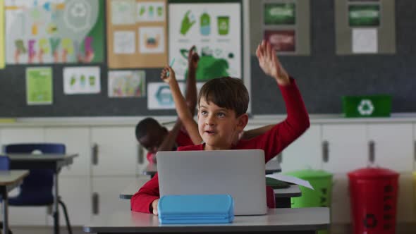 Portrait of happy caucasian schoolboy with laptop sitting raising hand to answer questions alt