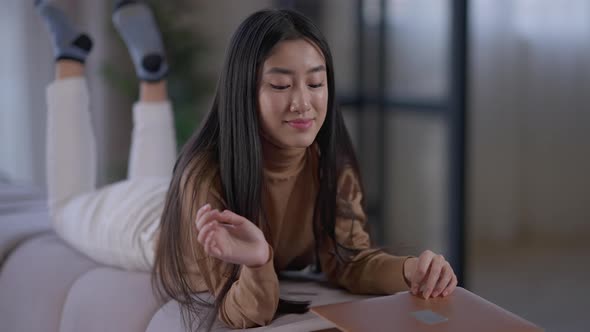Wide Shot of Happy Asian Freelancer Closing Laptop Lying on Couch and Smiling Looking at Camera alt