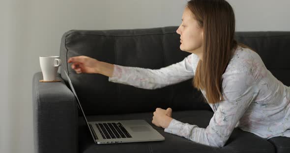 Pretty young woman on cofa, drinks coffee or tea and works on laptop at home. Girl works at home.