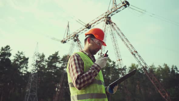 Electrical Equipment Worker Near High Voltage Tower Using Walkie Talkie and Clipboard alt