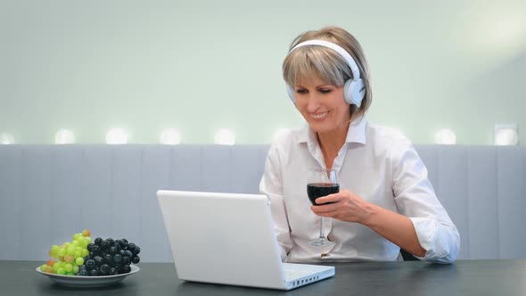 A Happy Elderly Woman with Headphones Drinking Wine Calls a Friend a Relative From Home on a Laptop