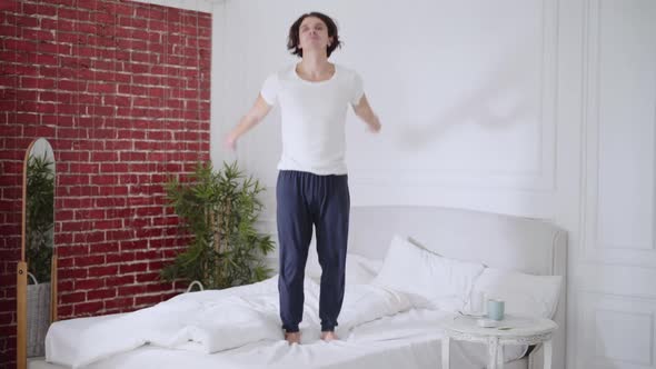 Wide Shot of Happy Carefree Young Man Jumping on Bed Smiling alt