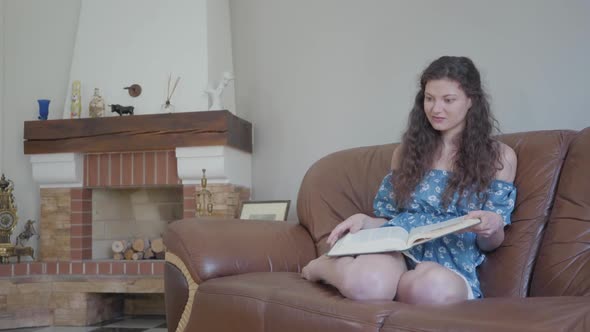 Portrait Adorable Curly Woman Sitting on the Leather Sofa at Home Reading Book alt