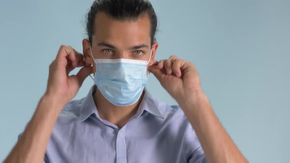 A Good-Looking Man Putting On A Surgical Mask Isolated On Light Blue Background - Covid-19 Outbreak alt