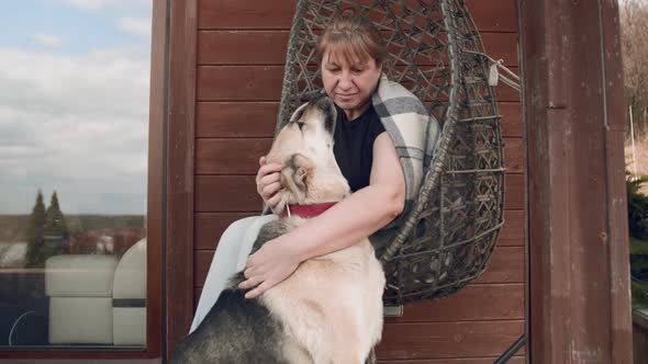 A woman plays and petting a shepherd dog while sitting in a wicker chair in a country house.