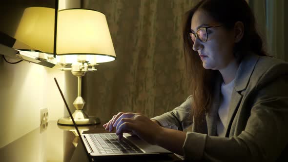 Young Woman in Glasses and a Suit is Typing Text on a Laptop Under the Light of a Table Lamp alt