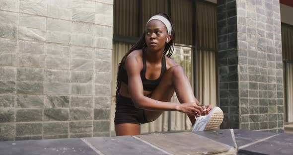 African american woman exercising outdoors wearing wireless earphones tying shoe in the city alt