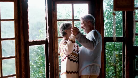Happy senior couple in love dance at home agains a big windows view with nature woods outside alt