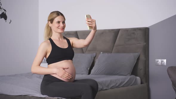 Young Caucasian Pregnant Woman Takes Pictures of Herself on Her Smartphone Poses Smiles