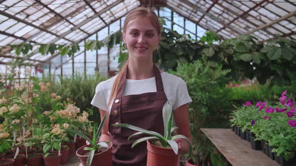 Young Blonde Girl Walks Through a Greenhouse with Pots of Flowers
