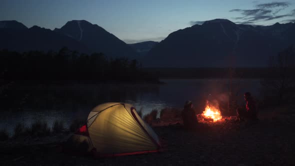 Two Tourists Sitting at Tent with Campfire and Looking at the Mountain