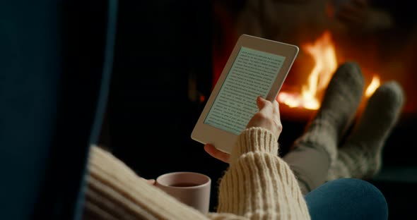 Woman Reads eBook on Electronic Reader By Fireplace in Cozy Room at Night