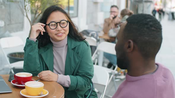 Cheerful Asian Woman Speaking with Afro Man in Outdoor Cafe alt