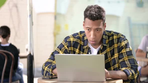 Young African Man Wondering in Shock on Laptop Outdoor Cafe alt