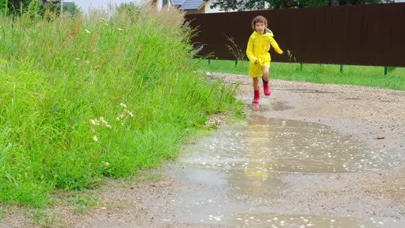 A girl in red rubber boots and a yellow raincoat runs through puddles after a rain in the village. S alt