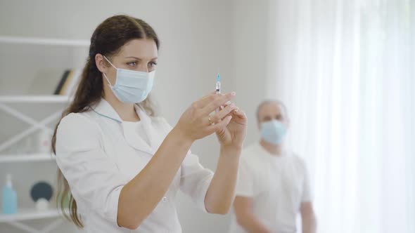 Serious Beautiful Woman in Face Mask Preparing Injection for Patient Sitting in Hospital Ward at the alt