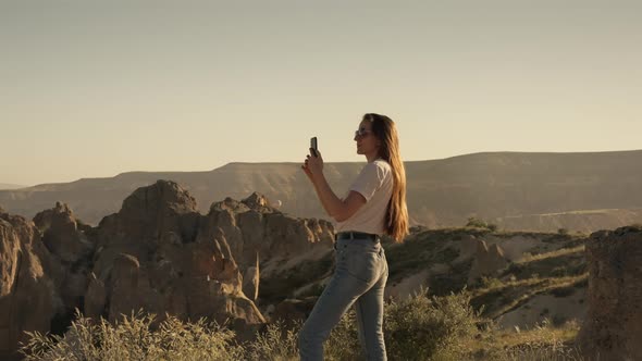Woman with Long Hair Takes Photo of Rocks and Mountains in Valley in Morning