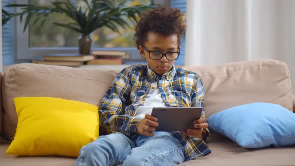 Cute African Child in Glasses Sitting on Sofa Playing Game on Digital Tablet. alt