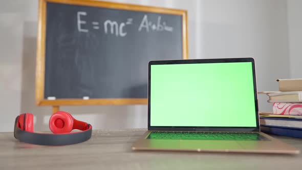 Closeup of Laptop with Chromakey Screen on Desk in Classroom with Headphones and Blurred Chalkboard alt