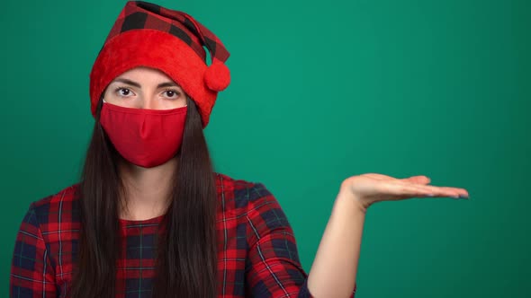 Young Woman in Santa Hat and Medical Red Mask Holds Invisible Object, Keeps Palm Raised alt