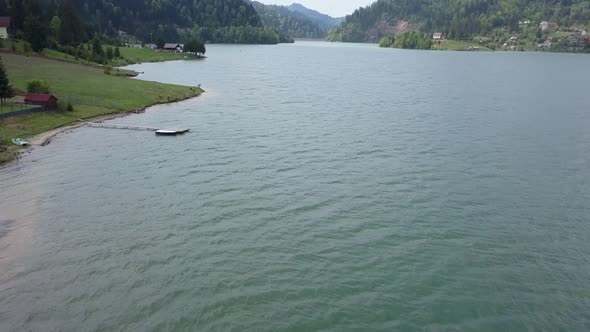 Aerial Panning Shot of edge of lake with a cabin and trees in the background and a pier in the foreg alt