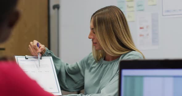 Caucasian businesswoman holding paperwork talking to colleague in office and smiling alt