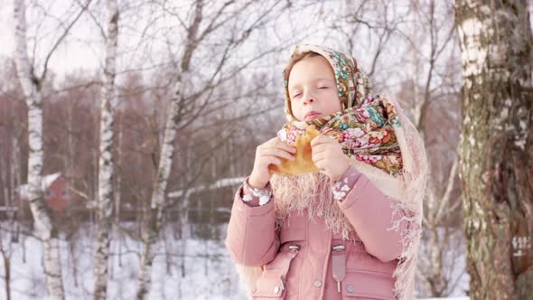 Cute girl in a traditional Russian headscarf with bagels eats pancake on winter background. alt