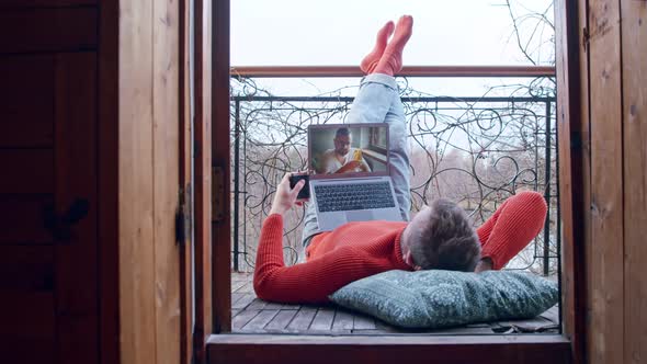 Man Is Lying on a Balcony Drinking From a Flask and Using a Video Call Talking To a Friend Who Is alt