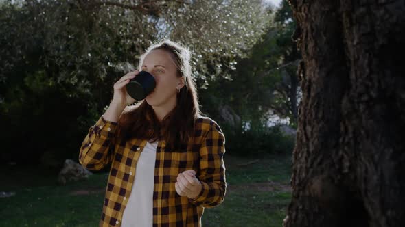 Young woman in a yellow shirt drinks hot tea from thermos in park on Sunny day. alt