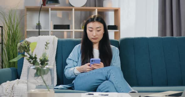 Asian Girl with Black Hair in Casual Clothes Sitting on Soft Couch at Home and Texting on Mobile alt