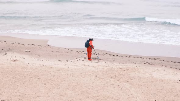 Man in Red Jumpsuit Looking for Lost Luxuries with Electronic Metal Detector on the Beach on a alt