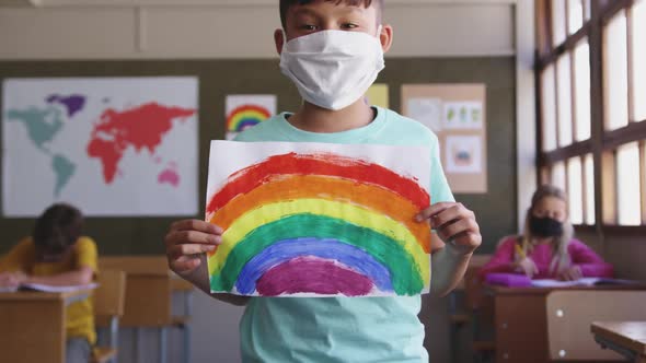 Boy wearing face mask holding a rainbow painting in class at school  alt