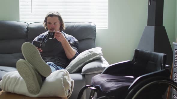 Happy caucasian disabled man sitting on sofa in living room using smartphone and drinking coffee alt