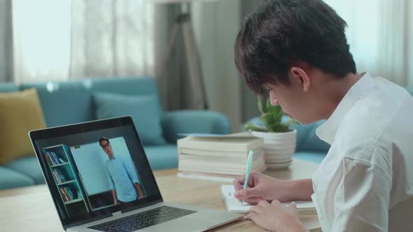Asian Man Student Look At Teacher In Computer Screen And Write In The Notebook While Studying Online alt