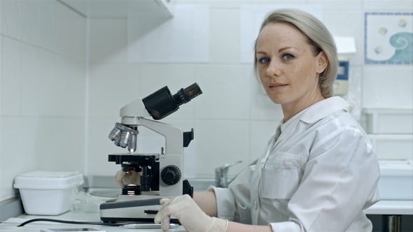 Pretty female with microscope looking at camera in laboratory