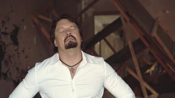 Portrait of a Brutal Young Man with a Beard Stands in a Shattered Room and Kneads His Neck Closeup alt