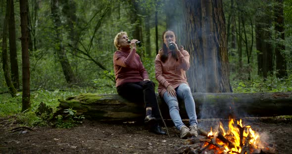 An Elderly Woman and a Middle-aged Woman Are in the Forest and They Are Sitting Near a Burning Fire alt