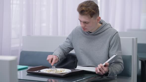 Hurrying Busy Caucasian Young Man Reading Book Eating Dessert in Canteen Indoors alt