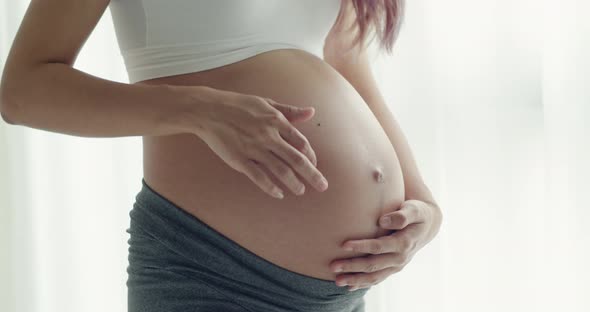 Asian happy pregnant woman is standing beside windows and touching her belly.