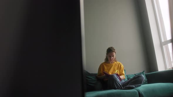 The Girl Makes Notes in a Notebook Sitting on a Bright Sofa alt