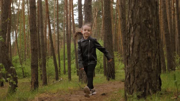 The Little Girl Runs Along the Path Road in the Pine Forest. Dressed in the Leather Jacket.