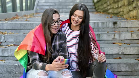 Beautiful Lesbian Couple with Rainbow Flag Using a Mobile Phone in the Street