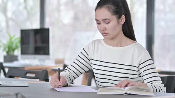 Young Asian Woman Doing Paperwork with Book in Office alt