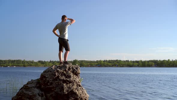 A Young Man Stands on a Stone Near the River Admiring the Landscape. A Man in Nature. alt