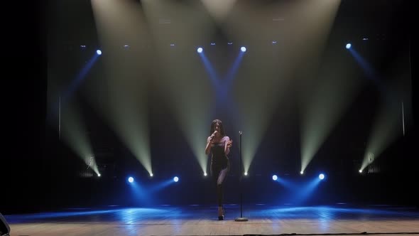 A Young Woman in a Black Suits Sings a Song and Dances on a Dark Stage. alt