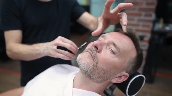 Barber master shaving handsome mature bearded man using straight razor in salon.
