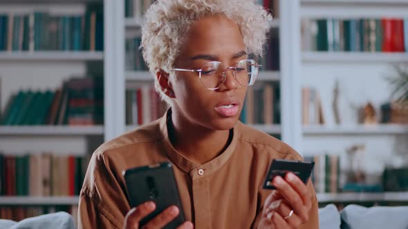Young African American Woman Makes Online Purchases in Mobile Phone Sits on Sofa alt